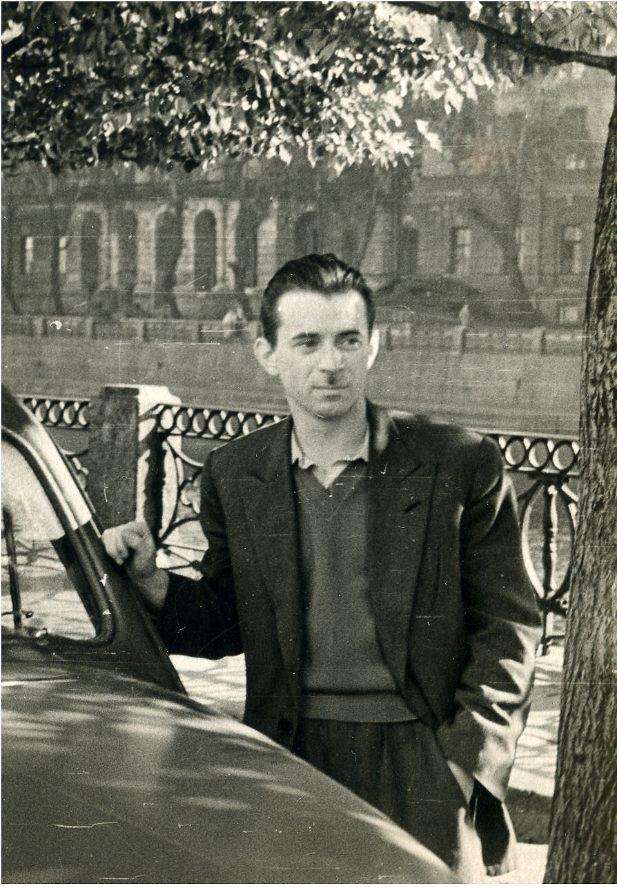 Черно-белое фото, мужчина в пиджаке стоит положив руку на автомобиль